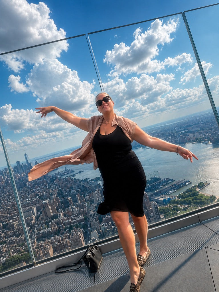 Woman posing with arms outstretched against a city skyline and blue sky with clouds.